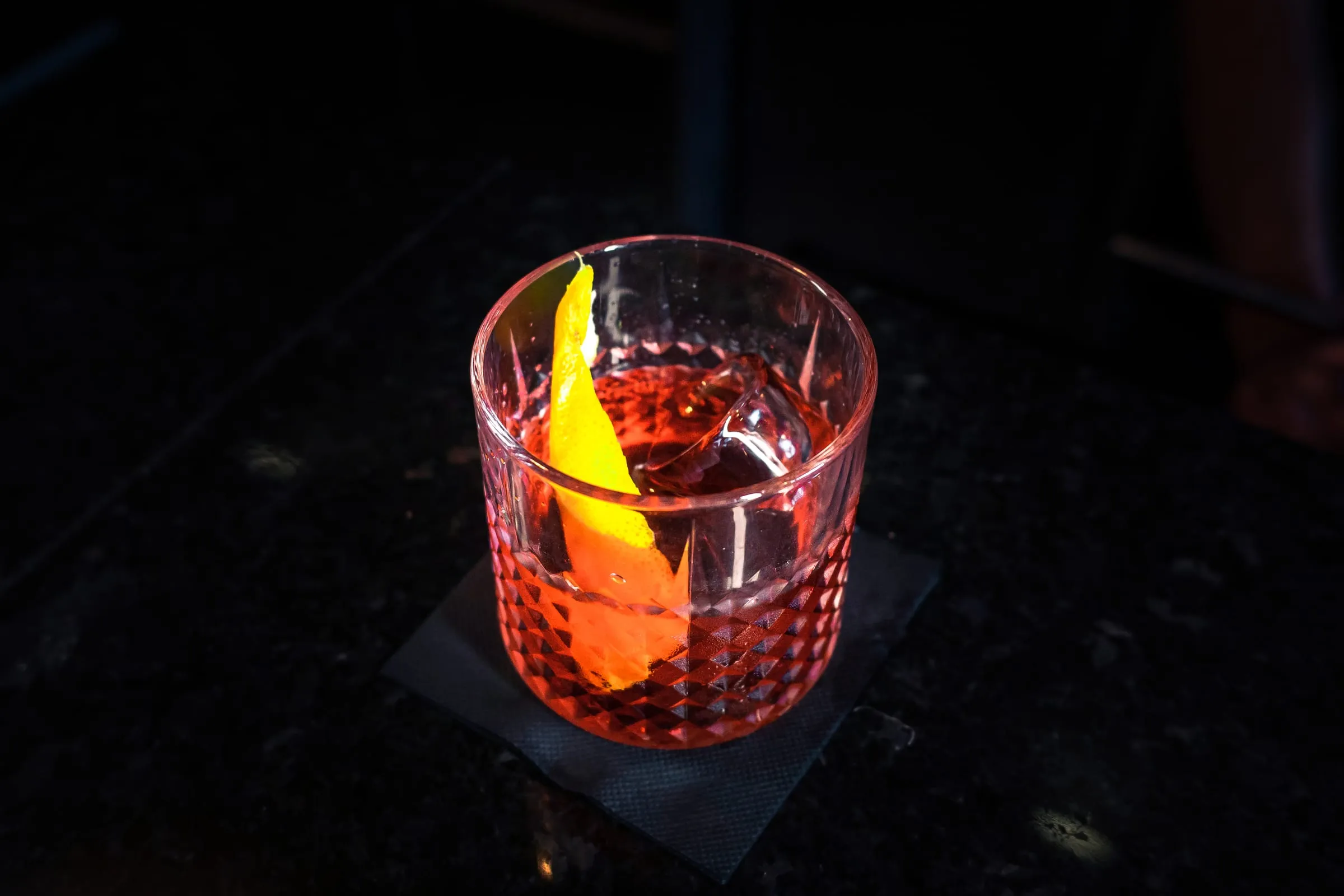 Copo de negroni com casca de laranja, iluminado no escuro.