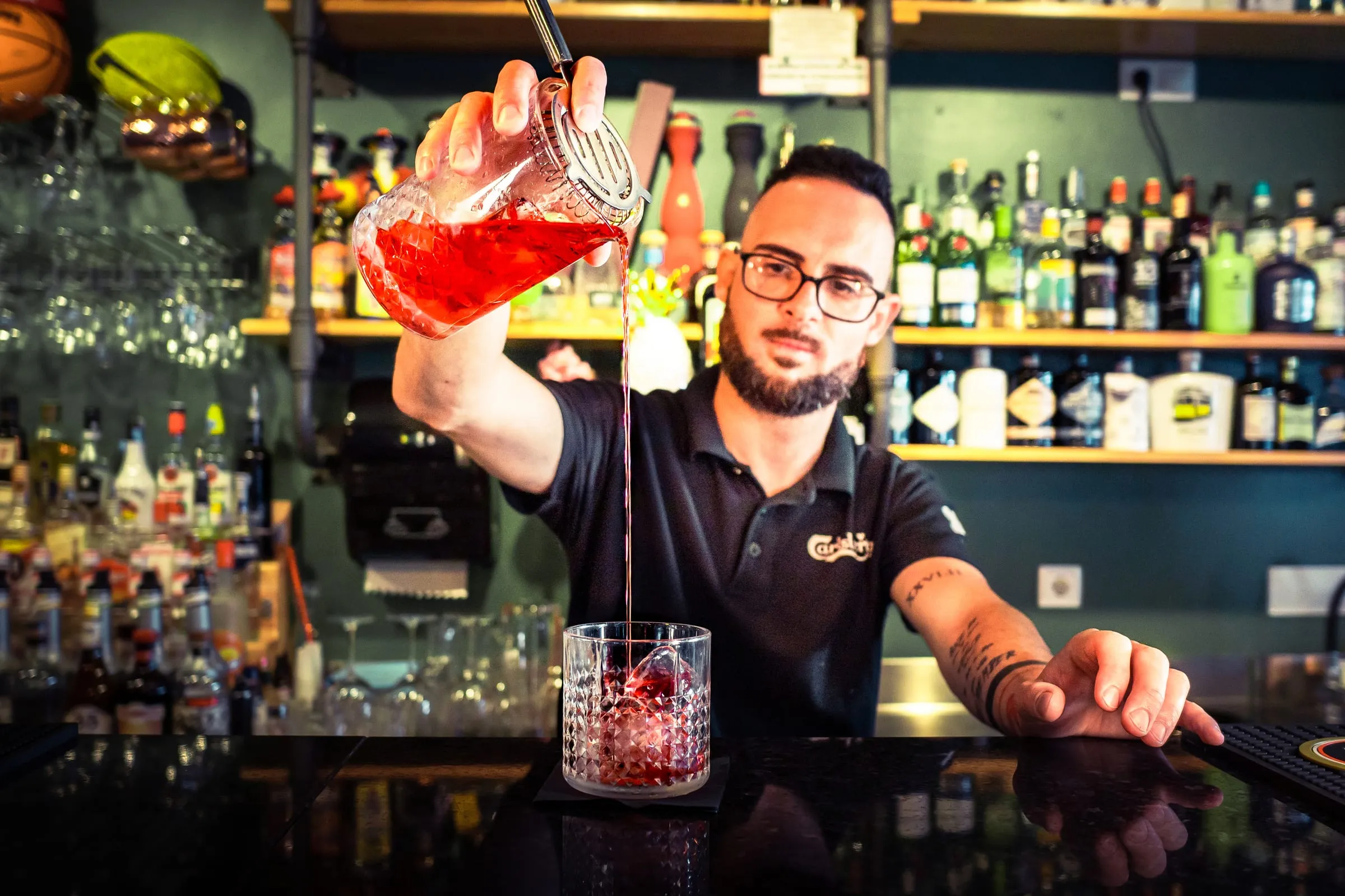 Barman coa um cocktail vermelho em altura à frente das prateleiras de garrafas.
